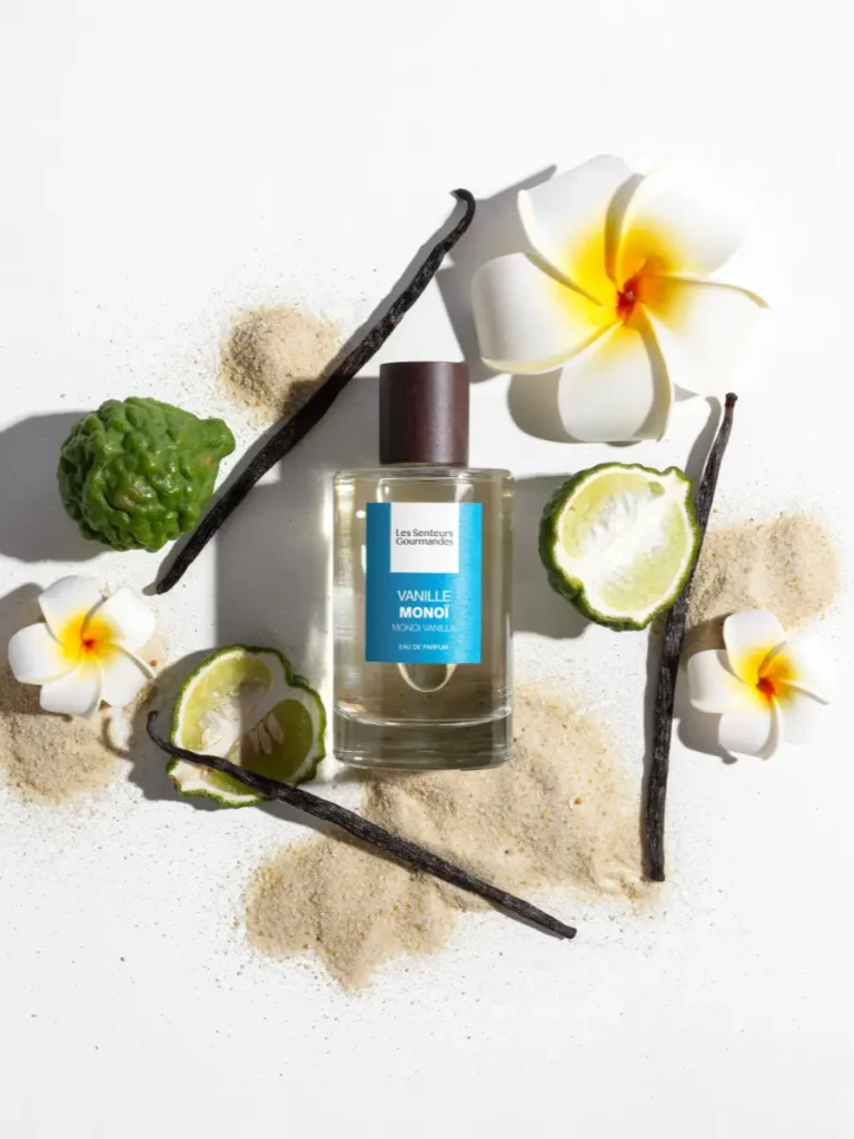 Flacon de parfum Vanille Monoi entouré de fleurs de tiaré, gousses de vanille, citron vert et sable clair.